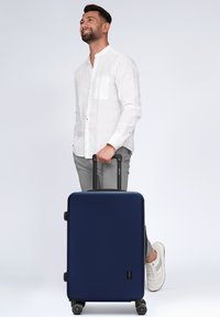 Marineblauwe hardshellkoffer met een gladde textuur, uitschuifbare handvat en vier wielen voor mobiliteit, geplaatst naast een persoon in lichte kleding.
