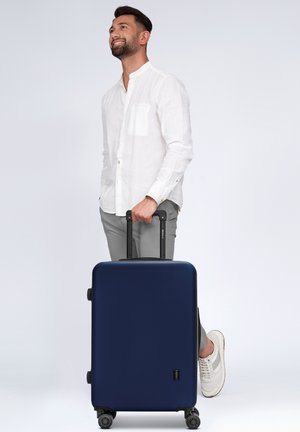 Valigia rigida blu navy con superficie liscia, manico retrattile e quattro ruote per la mobilità, posizionata accanto a una persona in abbigliamento leggero.