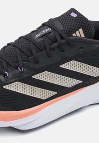 Chaussure de course noire Adidas avec des bandes argentées perforées, des lacets noirs, des accents violets et une semelle intermédiaire couleur pêche avec le texte "Lightstrike Pro".