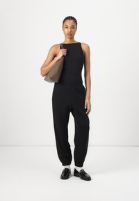 Black ribbed tank top paired with black tapered joggers. Model holds a brown leather bag. Shoes are shiny black loafers with white socks.