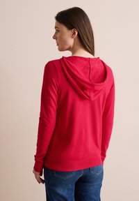 Röd hoodie av mjukt tyg, med en rymlig huva, ribbade manschetter och fåll, samt en enkel design. Bärs tillsammans med denimjeans.