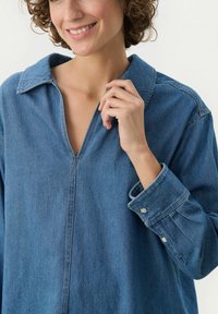 Camicia di denim di blu medio, con scollatura a V, maniche lunghe e polsini con bottone. Texture morbida con dettagli di cucitura discreti.