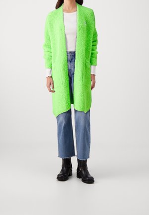Personne portant un cardigan texturé vert vif sur un haut blanc, un jean court bleu et des bottines noires, debout devant un fond uni.
