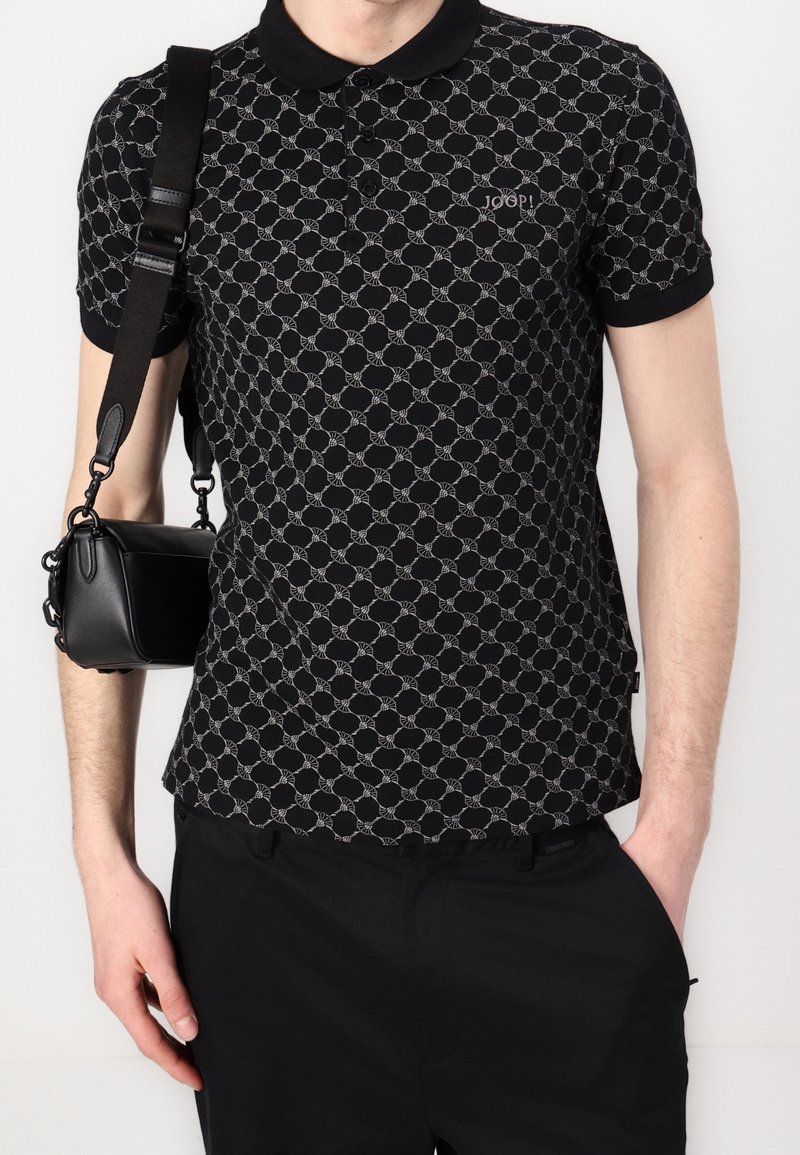 Homme portant un polo noir à motifs avec le logo "Joop!", portant un petit sac à bandoulière noir avec une sangle large.