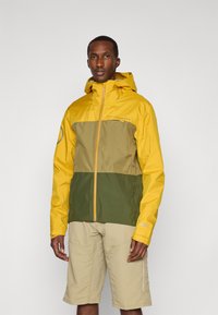 Wasserabweisende Jacke in Gelb und Olivgrün, mit einem Halb-Reißverschluss-Design, verstellbarer Kapuze und leichtem Stoff. Getragen mit beigen Shorts.