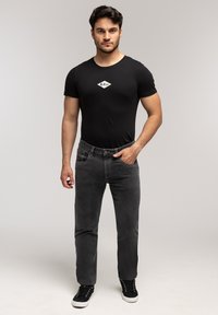 Mężczyzna stojący w obcisłej czarnej koszulce z logo, ciemnoszarych dżinsach i czarnych tenisówkach, z jedną ręką w kieszeni.