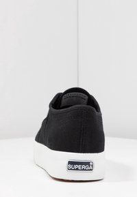 Svart canvas sneakers med låg design, vit gummisula och en svart etikett på häl som visar varumärket "SUPERGA."