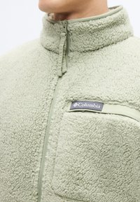 Fleecejacke in Hellgrün mit hohem Kragen, Reißverschluss und Brusttasche, die ein schwarzes Columbia-Logo zeigt. Weiches, strukturiertes Material.