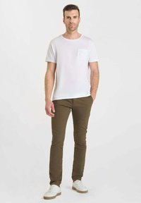 Man die naar voren staat en een wit T-shirt met korte mouwen, olijfgroene broek en witte sneakers met gumzolen draagt.