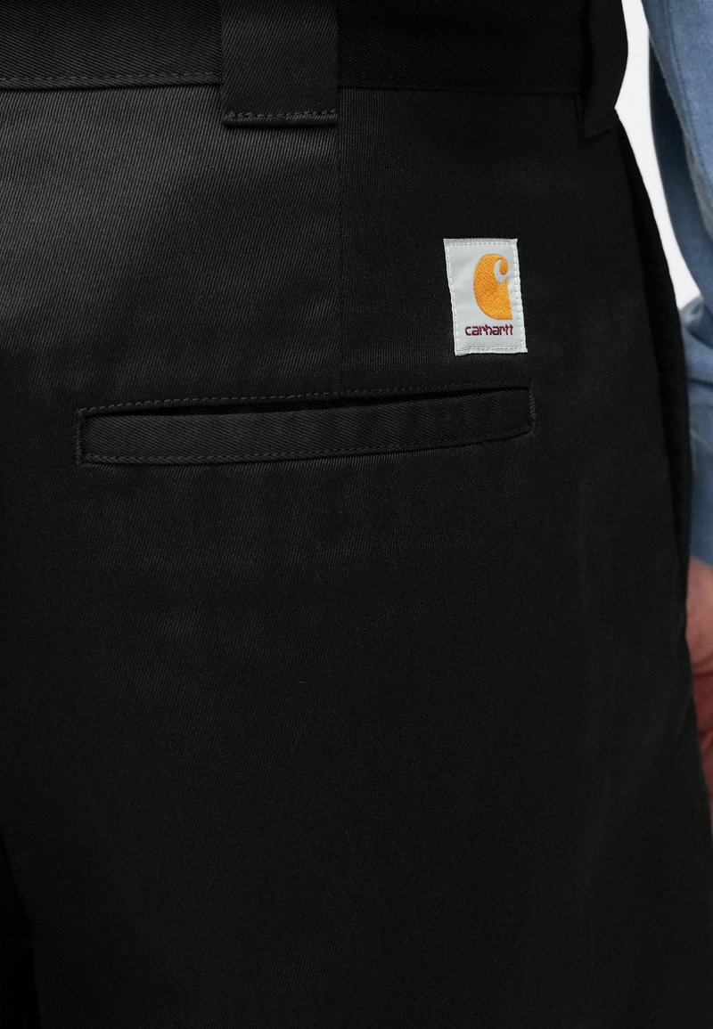 Pantaloni da lavoro neri con una texture liscia, caratterizzati da una piccola etichetta con il logo Carhartt arancione su sfondo bianco e una tasca posteriore.