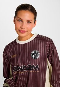 Braun gestreiftes Sporttrikot mit langen Ärmeln und beigen Akzenten, mit einem Logo auf der Brust und Markentext in Gold auf der Vorderseite.