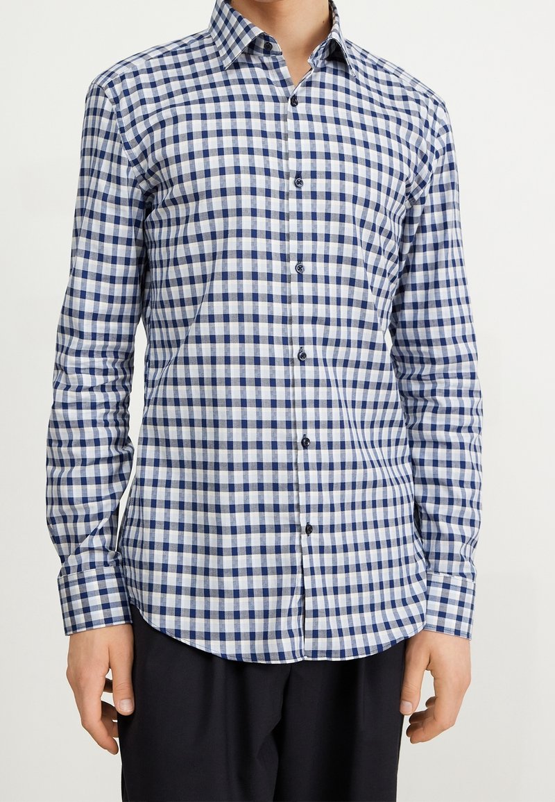 Uomo che indossa una camicia a maniche lunghe a quadri blu e bianchi con pantaloni neri su uno sfondo neutro.