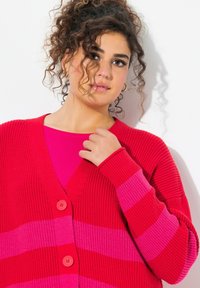 Cardigan rayé rouge et rose en tissu tricoté texturé, avec un décolleté en V profond et deux grands boutons à l'avant.