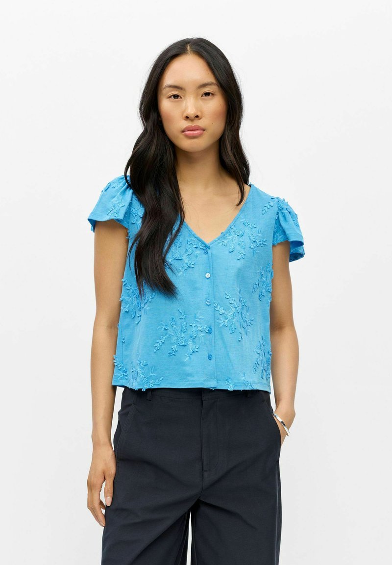 Femme aux longs cheveux foncés portant un chemisier bleu brodé à manches courtes et un pantalon noir, debout avec une main dans la poche sur fond blanc.