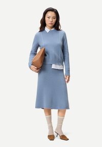 Kvinna i blå stickad tröja och kjol som håller en stor brun väska, iklädd beige strumpor och bruna klackskor på vit bakgrund.