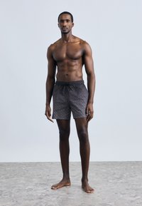 Črne kopalne hlače z vzorcem, elastičnim pasom in vrvico, narejene iz lahkega materiala, model stoji boso na teksturirani površini.