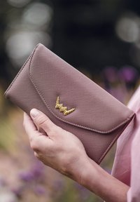 Pink textured wallet with a flap closure, featuring a gold wave-shaped accent on the front. Held in a hand against a blurred background.
