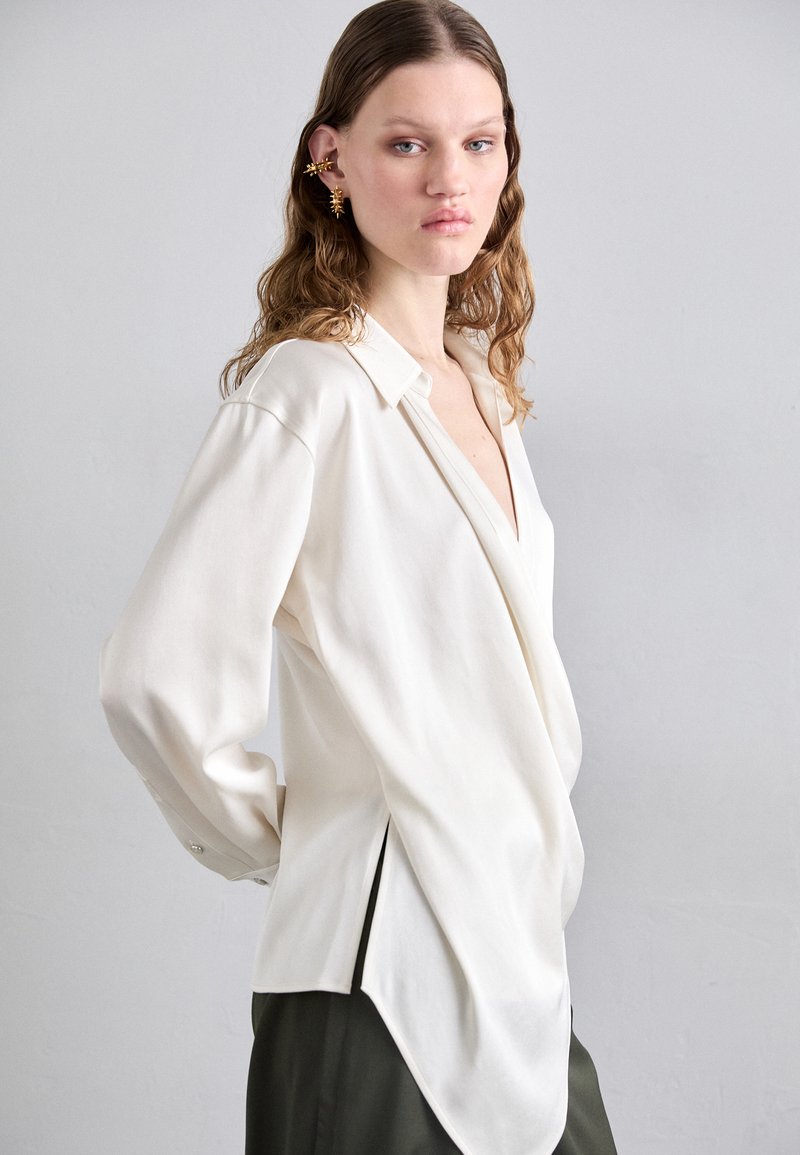 White long-sleeve shirt with a deep V-neck, side slits, and a relaxed fit. Features a buttoned cuff and smooth texture. Styled over dark pants.