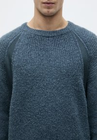 Dunkelblauer Pullover aus einem strukturierten, flauschigen Material. Verfügt über einen gerippten Ausschnitt und seitliche Akzente aus einem glatten, kontrastierenden Stoff.
