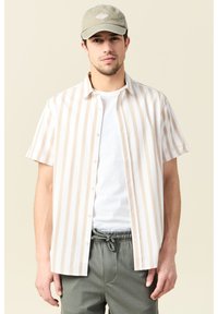 Camisa de botones de manga corta en blanco con rayas verticales marrón claro; tela suave; llevada sobre una camiseta blanca; combinada con pantalones verde oliva.