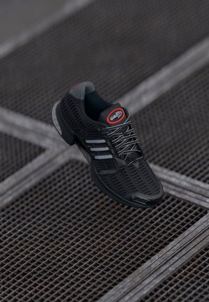 Chaussure de sport noire avec des bandes blanches et un logo rouge sur la languette, présentée au-dessus d'une surface en grille métallique.
