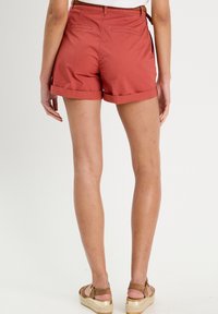 Shorts en coton de couleur rouille avec un ourlet retroussé, deux poches arrière et une ceinture tissée en accent. Portés avec des sandales à talons compensés marron.
