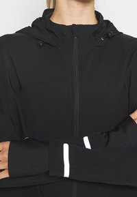 Donna che indossa una giacca nera con zip e cappuccio, con le braccia incrociate e strisce bianche riflettenti sui polsini delle maniche.