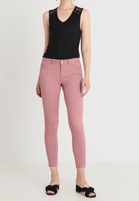 Kvinna i ärmlös svart top med spetsdetalj, figurnära dammrosa jeans och svarta öppna klacksandaler står mot vit bakgrund.