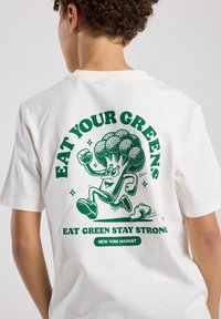 White cotton t-shirt featuring green graphic of a running broccoli character with text: "EAT YOUR GREENS" and "EAT GREEN STAY STRONG."