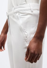 Πλάγια όψη ενός ατόμου που φοράει λευκό παντελόνι με κοντινή λήψη στη μέση της μέσης, στη λεπτομέρεια της τσέπης και το δεξί χέρι που ακουμπά δίπλα στο πόδι.