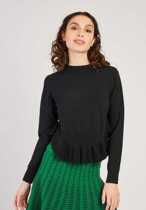 Pull en maille noire à manches longues, avec un col côtelé et un ourlet peplum. Porté par-dessus une jupe verte texturée présentant un motif rayé.