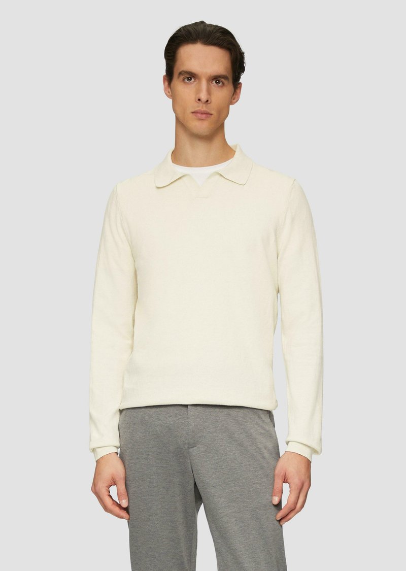 Jasno kremowa koszula polo z długim rękawem o miękkiej teksturze, z klasycznym kołnierzykiem i prążkowanymi mankietami. W połączeniu z szarymi spodniami.