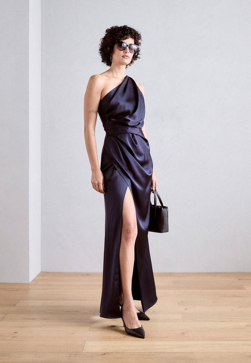 Granatowa satynowa sukienka z jednym ramieniem, z wysokim rozcięciem na udzie, paskiem w talii, w połączeniu z czarnymi szpilkami i małą torebką.