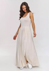 Długa, bezrękawna sukienka maxi w jasnym beżu z dopasowaną górą i swobodną, plisowaną spódnicą. Noszona z białymi sneakersami. Miękki, elastyczny materiał.