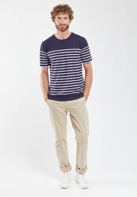 Marineblå stribet t-shirt med hvide vandrette striber, rund halsudskæring, parret med beige bukser og hvide sneakers, stående på en lys baggrund.