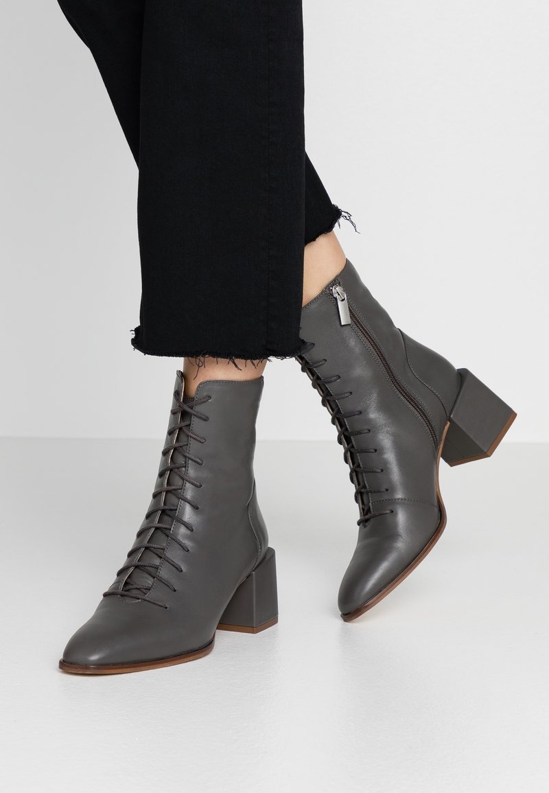 Femme portant des bottines gris foncé à lacets avec talons carrés et fermetures éclair sur les côtés, associées à un pantalon noir court aux ourlets effilochés.