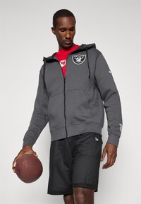 Szara bluza zapinana na zamek z kieszeniami kangurzymi, z logo Raiders. Nosi ją osoba włożona w czerwoną koszulę i czarne szorty siatkowe, trzymając piłkę nożną.