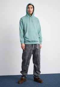 Felpa cappuccio di un azzurro chiaro con tasca a marsupio e cordini, abbinata a pantaloni cargo grigi testurizzati con tasche laterali. Scarpa marrone completa il look.