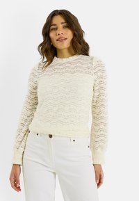 Top de manga larga de encaje en color blanco roto con patrones ondulados, cuello redondo y mangas texturizadas, combinado con vaqueros de color claro.