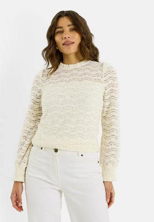 Top de manga larga de encaje en color blanco roto con patrones ondulados, cuello redondo y mangas texturizadas, combinado con vaqueros de color claro.