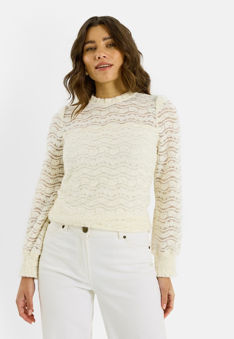 Top de manga larga de encaje en color blanco roto con patrones ondulados, cuello redondo y mangas texturizadas, combinado con vaqueros de color claro.