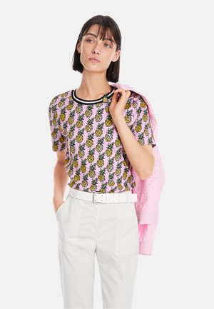 Short-sleeve pink t-shirt with a pineapple pattern, black neckline trim; paired with white trousers and a pink jacket draped over one shoulder.