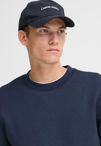 Joven con un sudadera azul marino y una gorra negra con el texto "Casual Apparel", mirando hacia un lado contra un fondo plano.