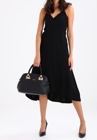 Zwarte wikkeljurk met een high-low zoom, gecombineerd met een gestructureerde zwarte handtas met gouden hardware en gevlochten handvatten. Zwarte hoge hakken.