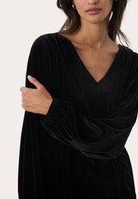 Black velvet dress with a V-neckline and long, slightly gathered sleeves. Soft texture and flowing shape emphasize elegance.