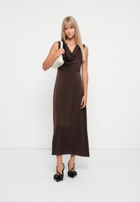Robe en satin marron avec un col drapé, corsage ajusté et silhouette fluide, accompagnée d'un sac à main blanc et de chaussures à talons pointus noires.