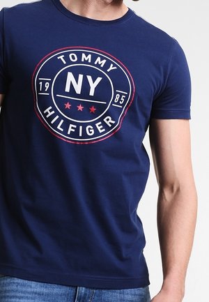Mężczyzna ubrany w granatowy t-shirt Tommy Hilfiger z okrągłym logo NY 1985 i czerwonymi gwiazdkami na klatce piersiowej.