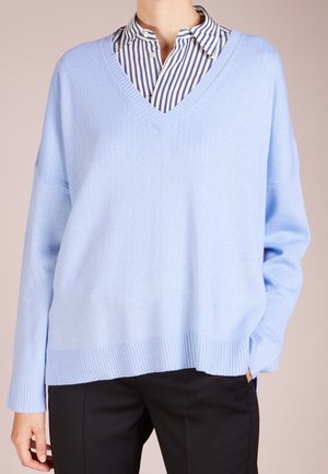 Jasnoniebieski, oversize sweter z dekoltem w serek wykonany z miękkiego materiału dzianinowego. Ma ściśnięte mankiety i dół, noszony na białej i niebieskiej koszuli w paski.