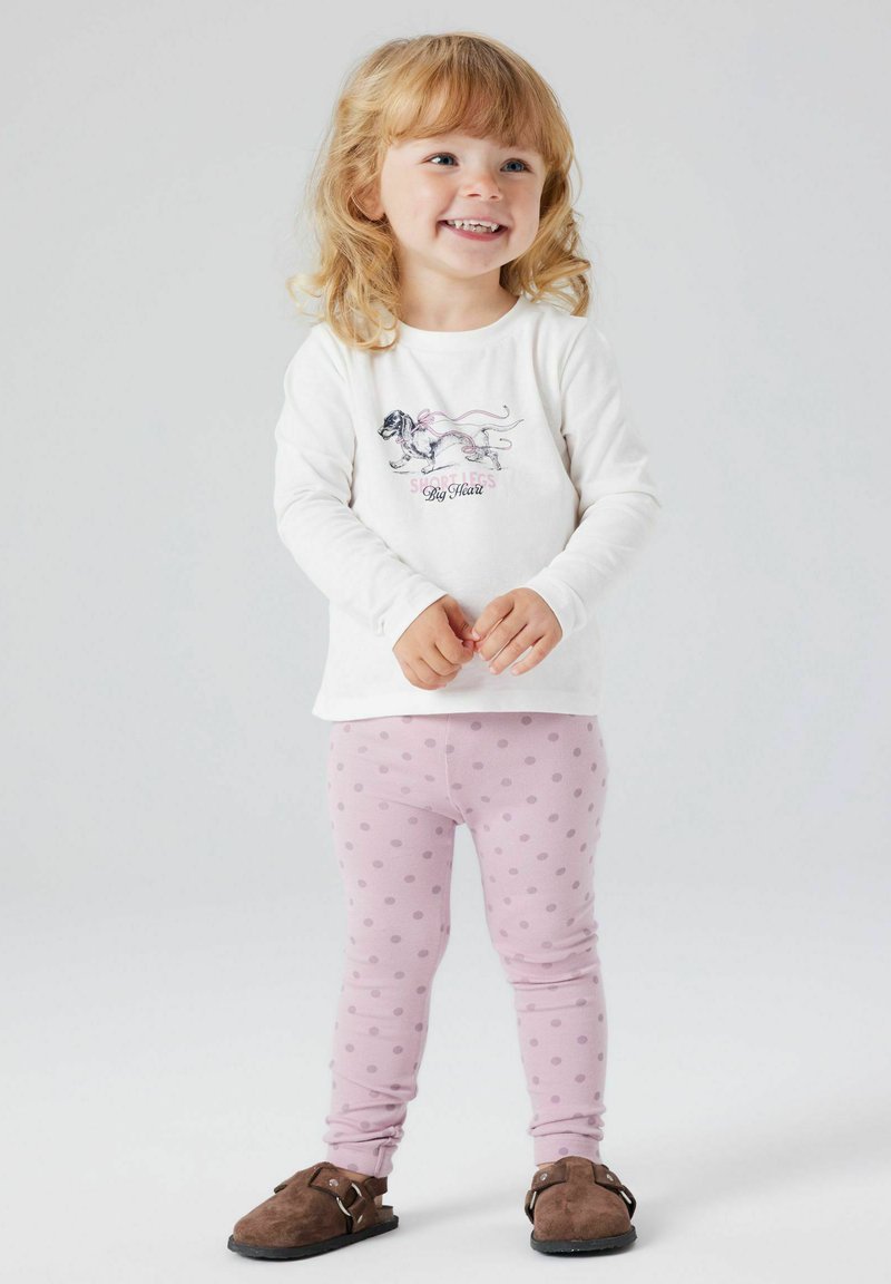 Hvid langærmet skjorte med hundetryk, pink tekst; pink leggings med mørkere prikker; brune slip-on sko med spænde detaljer.