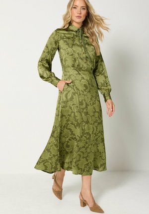 Femme blonde portant une robe midi fleurie à manches longues couleur vert olive avec un col à nœud et des chaussures à talons blocs couleur beige, debout sur un fond blanc.